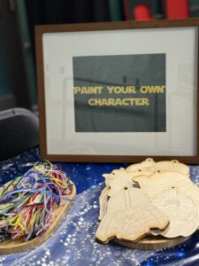 A crafting table displays a framed sign reading “Paint Your Own Character.” Wooden outlines of various Star Wars characters are stacked on trays next to bundles of colorful elastic strings.