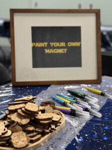 A framed sign reading “Paint Your Own Magnet” stands behind a tray of small laser-cut wooden Star Wars character magnets. Several water-brush style paint pens are lined up on a star-themed tablecloth.
