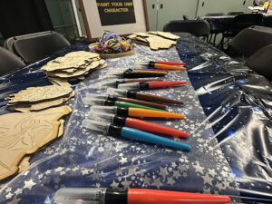 A long craft table covered with a starry blue tablecloth holds rows of colorful paint pens, stacks of wooden Star Wars character cutouts, and bundles of elastic cords, with chairs arranged around the table.