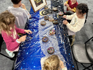Several children sit around a star-themed craft table, picking beads from bowls and assembling Star Wars–themed necklaces. The table is filled with different bowls of colorful beads and black necklace cords.