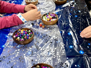 Close-up view of children’s hands reaching into bowls filled with colorful beads on a starry tablecloth, with necklace cords laid out nearby.