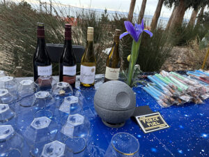 A table covered with a starry blue tablecloth displays several bottles of wine, upside-down plastic cups, a Death Star–shaped decoration, a single purple flower in a vase, and a tray of colorful lightsaber-themed treats.