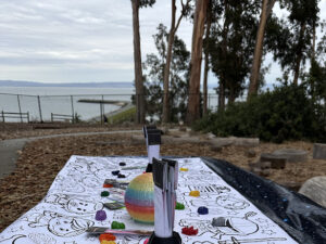 A picnic table covered with a Star Wars coloring tablecloth is set with markers and small rainbow decorations. Behind it, eucalyptus trees frame a scenic view of the bay.