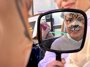 A child with detailed tiger face paint smiles while looking at their reflection in a handheld mirror. Part of the face painter’s face is visible in the background, along with colorful lights and decorations.