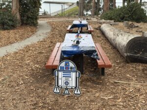 Two picnic tables outdoors are covered with Star Wars-themed tablecloths and small craft items, set among trees and wood chips. A large decorative cutout of R2-D2 is placed at the end of the nearest table.