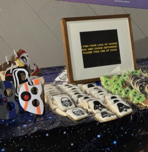 A table covered with a starry cloth displays felt Star Wars–themed masks, individually wrapped decorated cookies shaped like helmets and characters, and a framed sign that reads, “I find your lack of goodie bag and cookie disturbing. Please take one of each!”